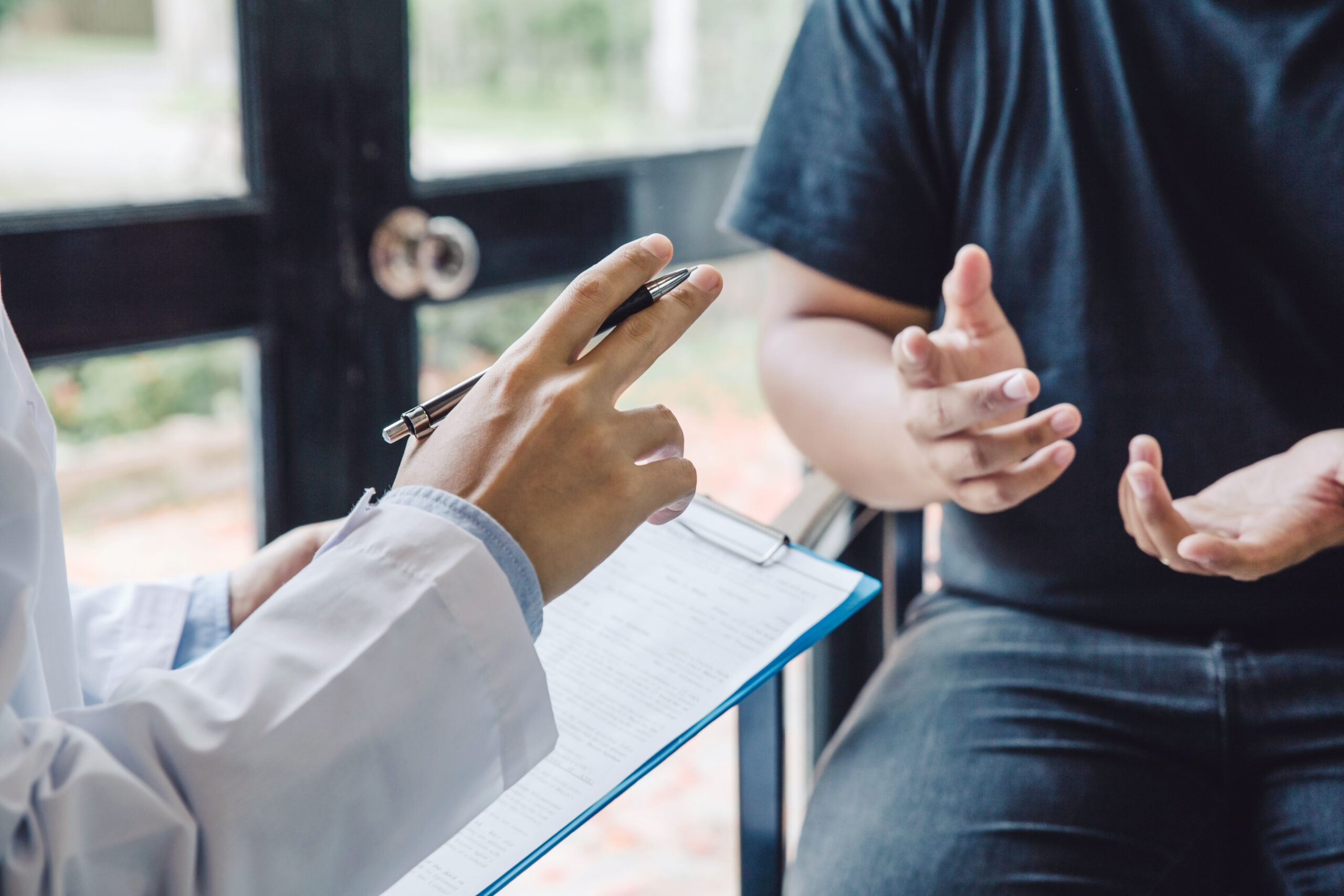 A set of hand (the doctor's) hold a pen in one hand and a clipboard in the other. Across from the doctor is another set of hands (the patient) making an open palm gesture. 