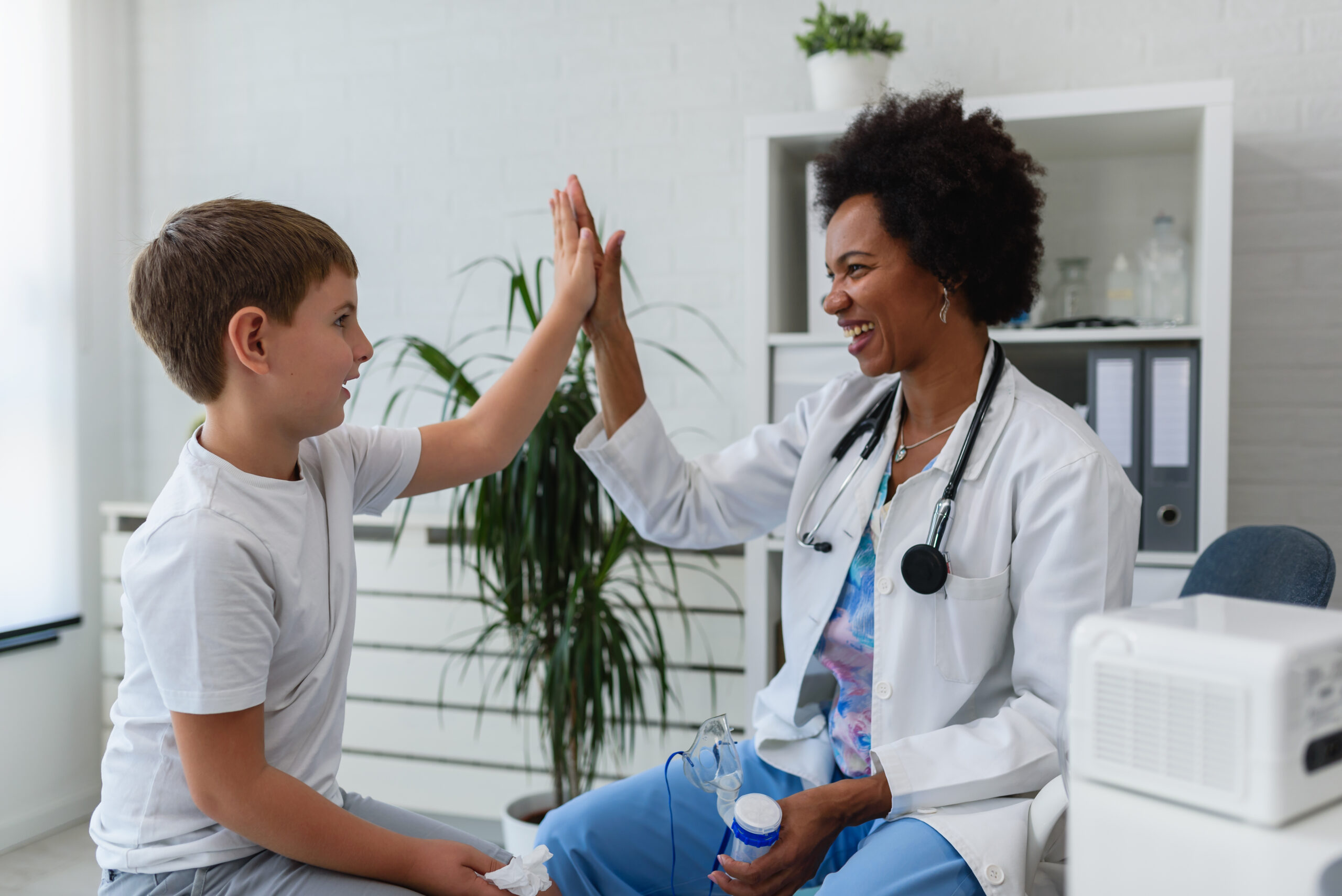 Smiling female doctor high-fiving a child patient. Both are seated in an exam room.